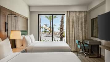 Une chambre d’hôtel équipée d’un lit, d’un bureau, d’une chaise, d’une télévision et offrant une vue sur une piscine et des palmiers.