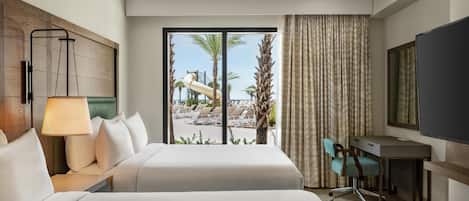 A hotel room with a bed, a desk, a chair, a TV, and a view of a pool and palm trees.