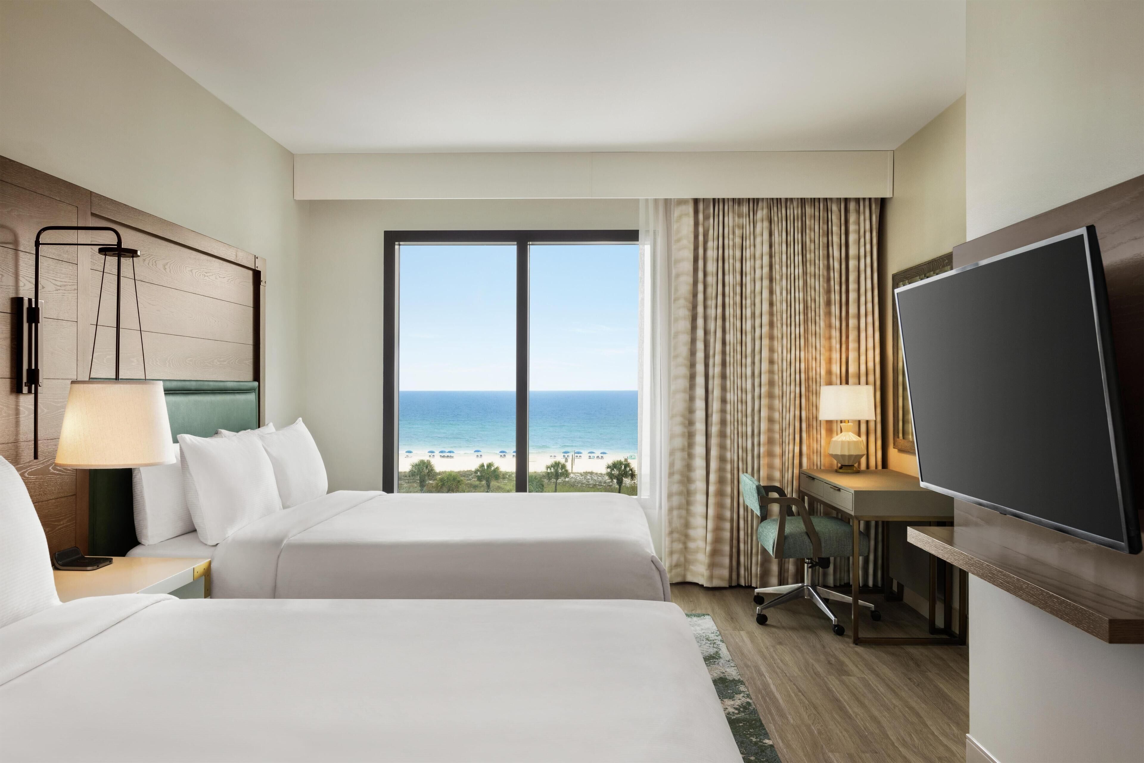 Habitación de hotel con dos camas, un escritorio, una televisión y vista a la playa.