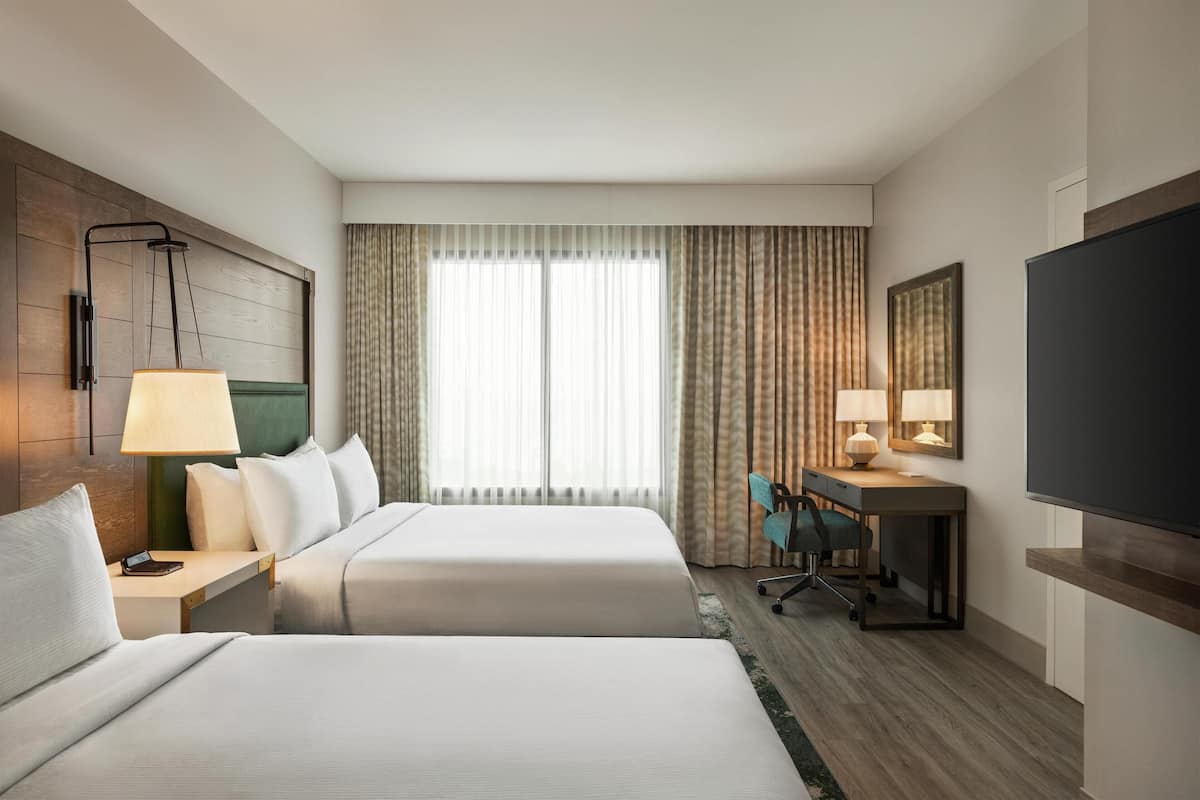 A hotel room with two beds, a desk with a chair, a TV, and a large window with curtains.