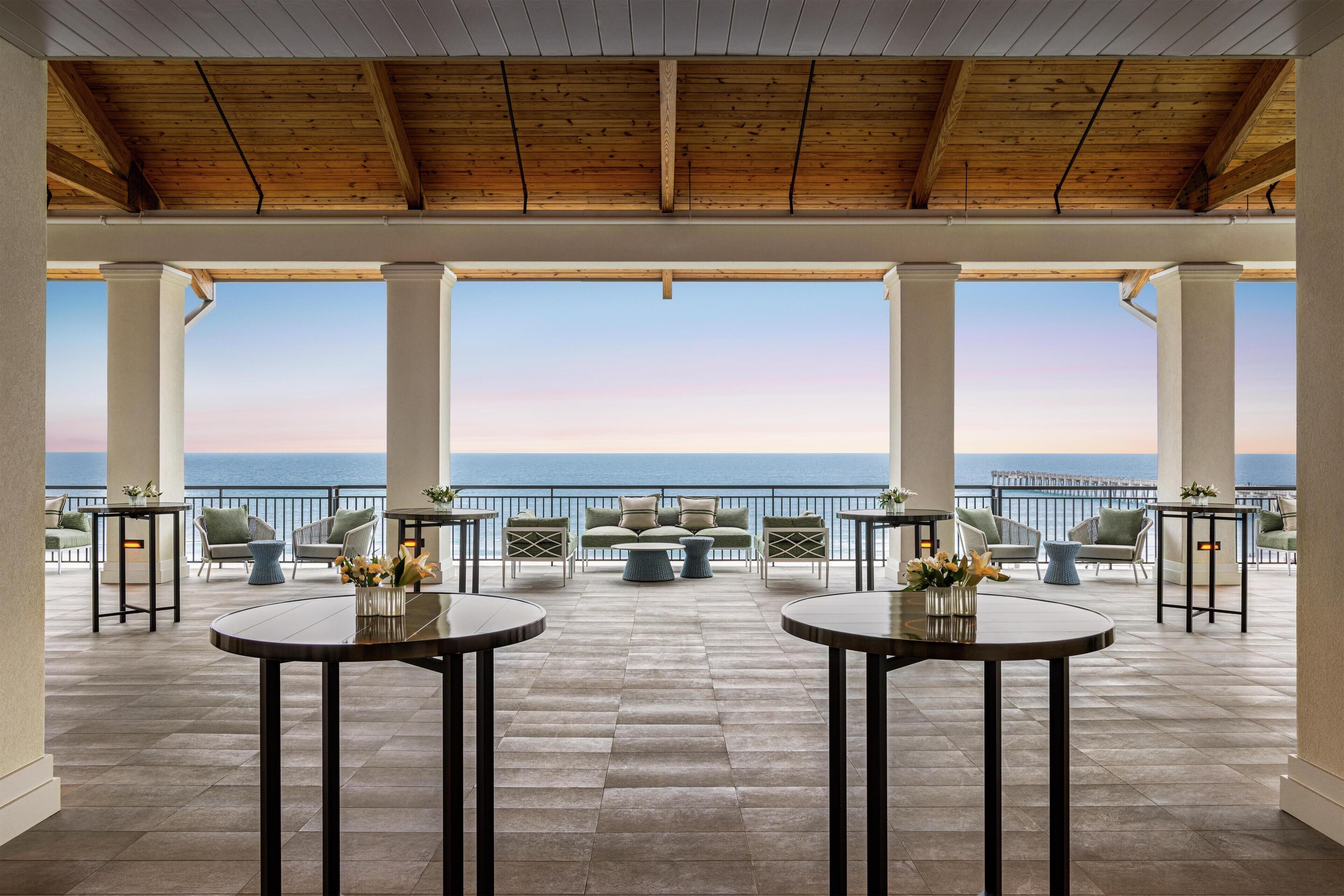A terrace with round tables and chairs, a view of the sea, and a wooden ceiling.