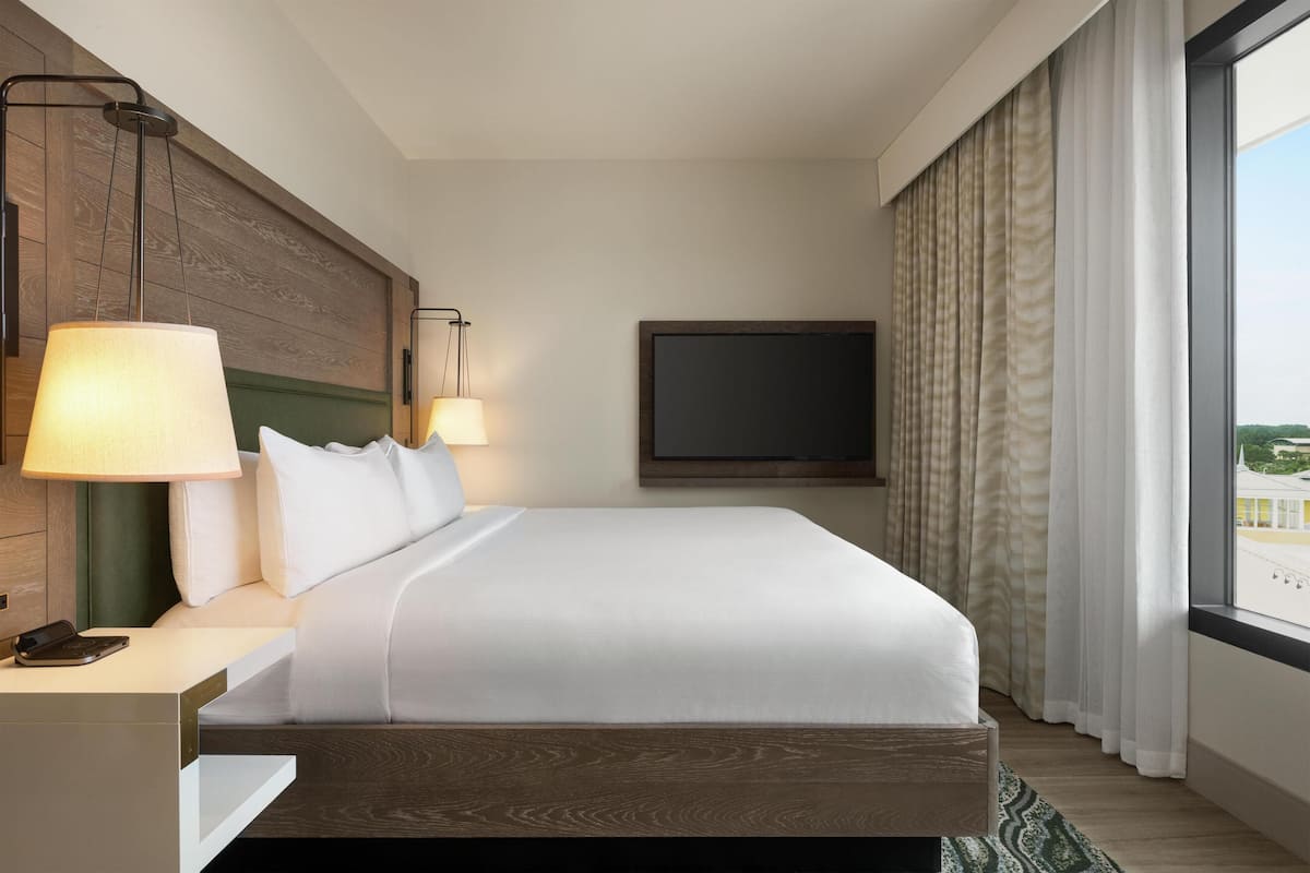A hotel room with a large bed, bedside lamps, a flat-screen TV, and a view of the city through large windows.