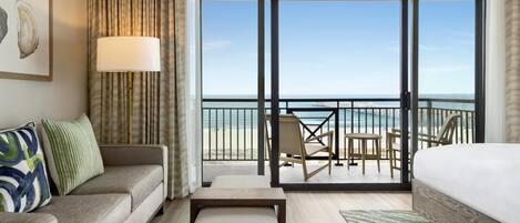 A hotel room with a balcony, a bed, a sofa, a table, and a view of the beach.