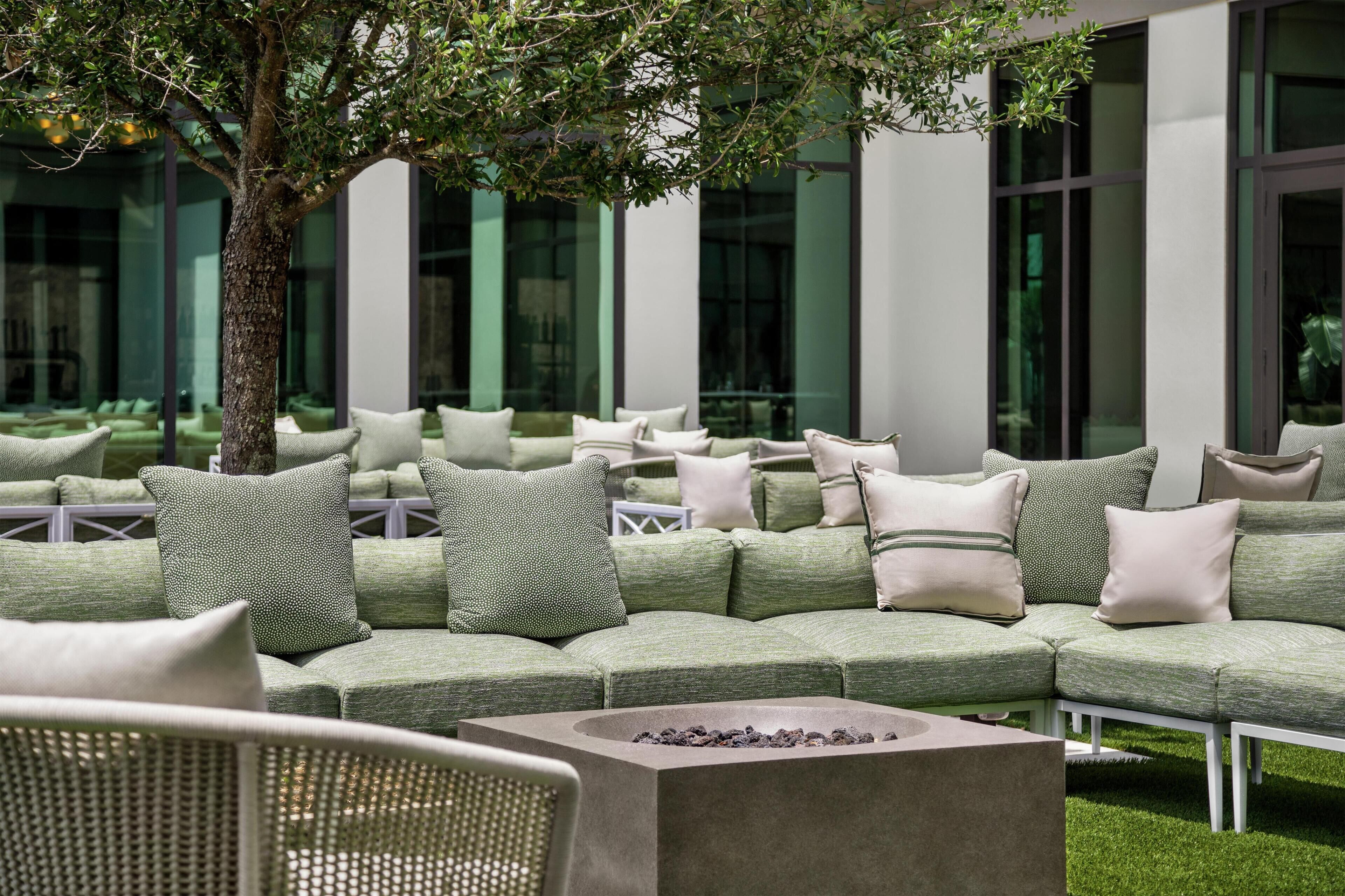 A modern outdoor seating area with a fire pit, cushioned seating, and a tree providing shade.