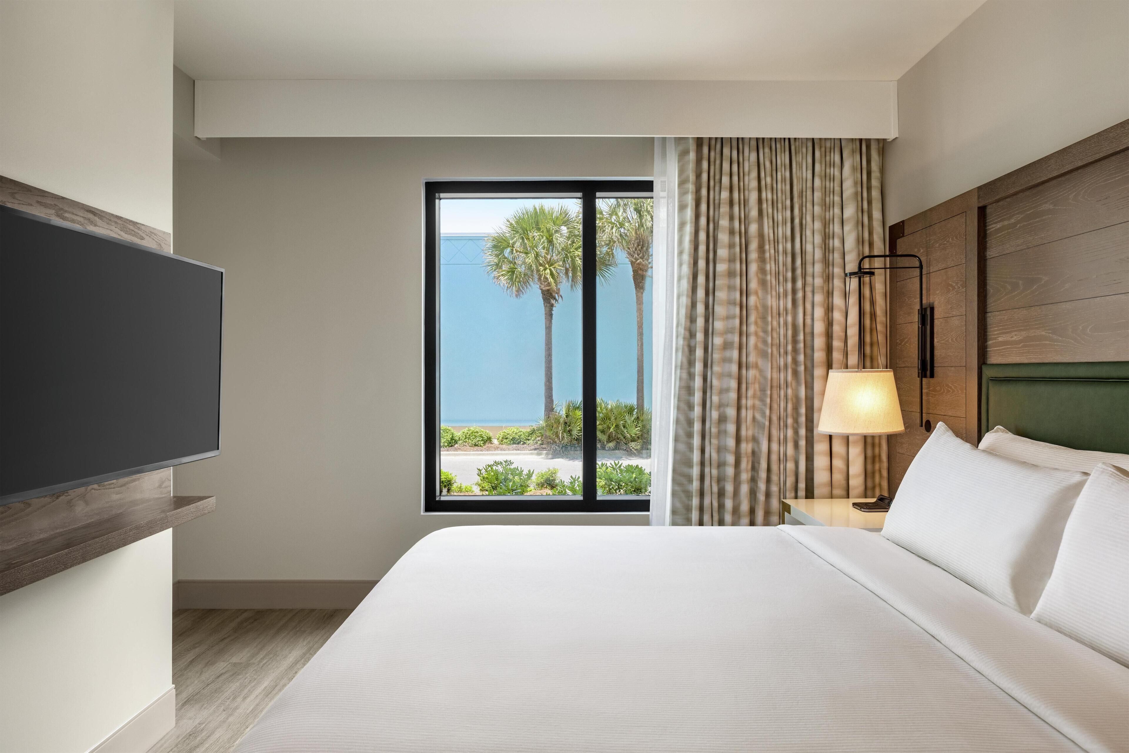 Una habitación de hotel con una cama grande, un televisor montado en la pared, una ventana con vistas a palmeras y al océano, y una lámpara de noche.