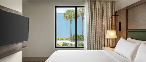 A hotel room with a large bed, a TV mounted on the wall, a window with a view of palm trees and the ocean, and a bedside lamp.