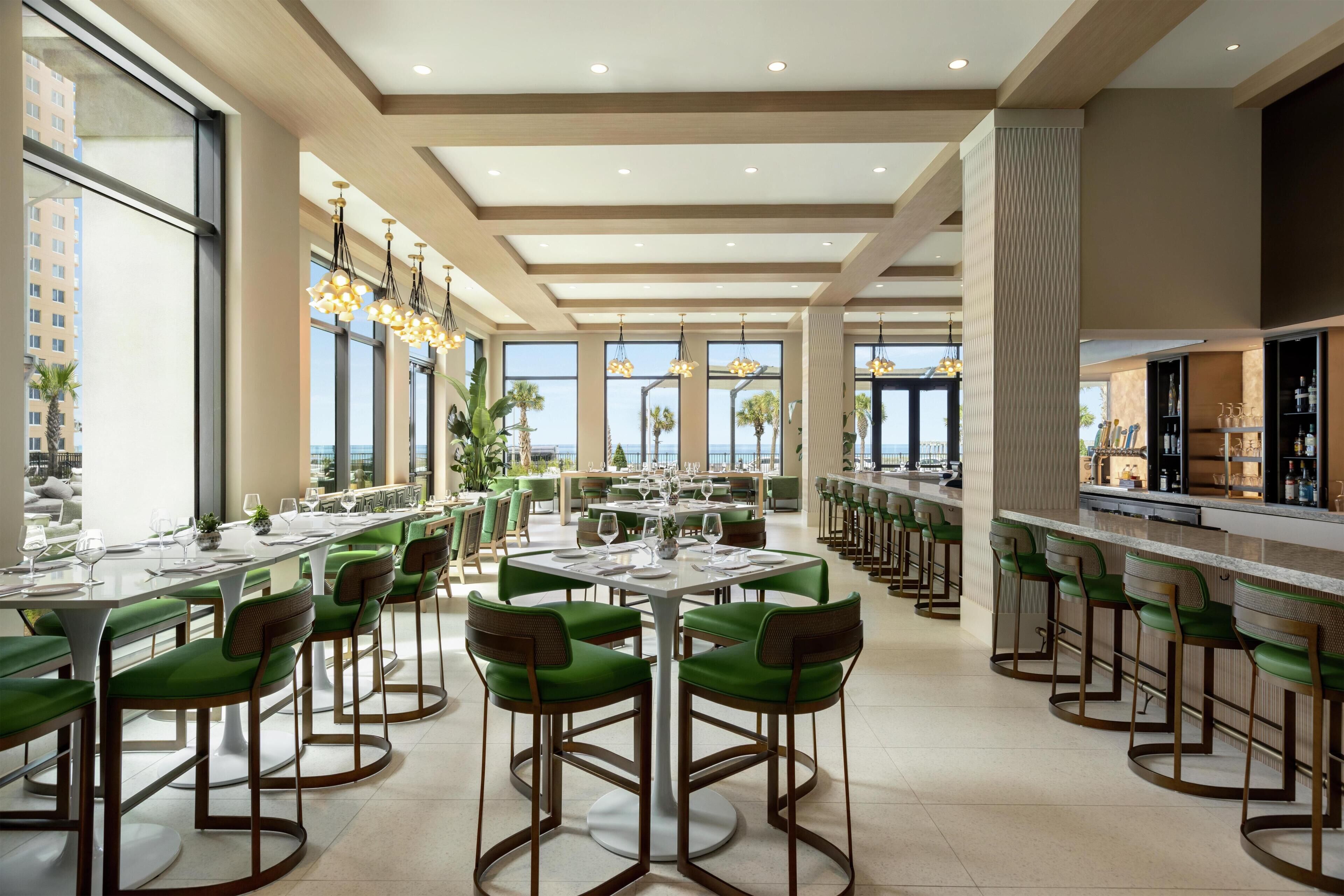 Un comedor amplio con mesas redondas y sillas verdes, ventanas grandes que ofrecen vistas de la ciudad, y una zona de bar con taburetes.