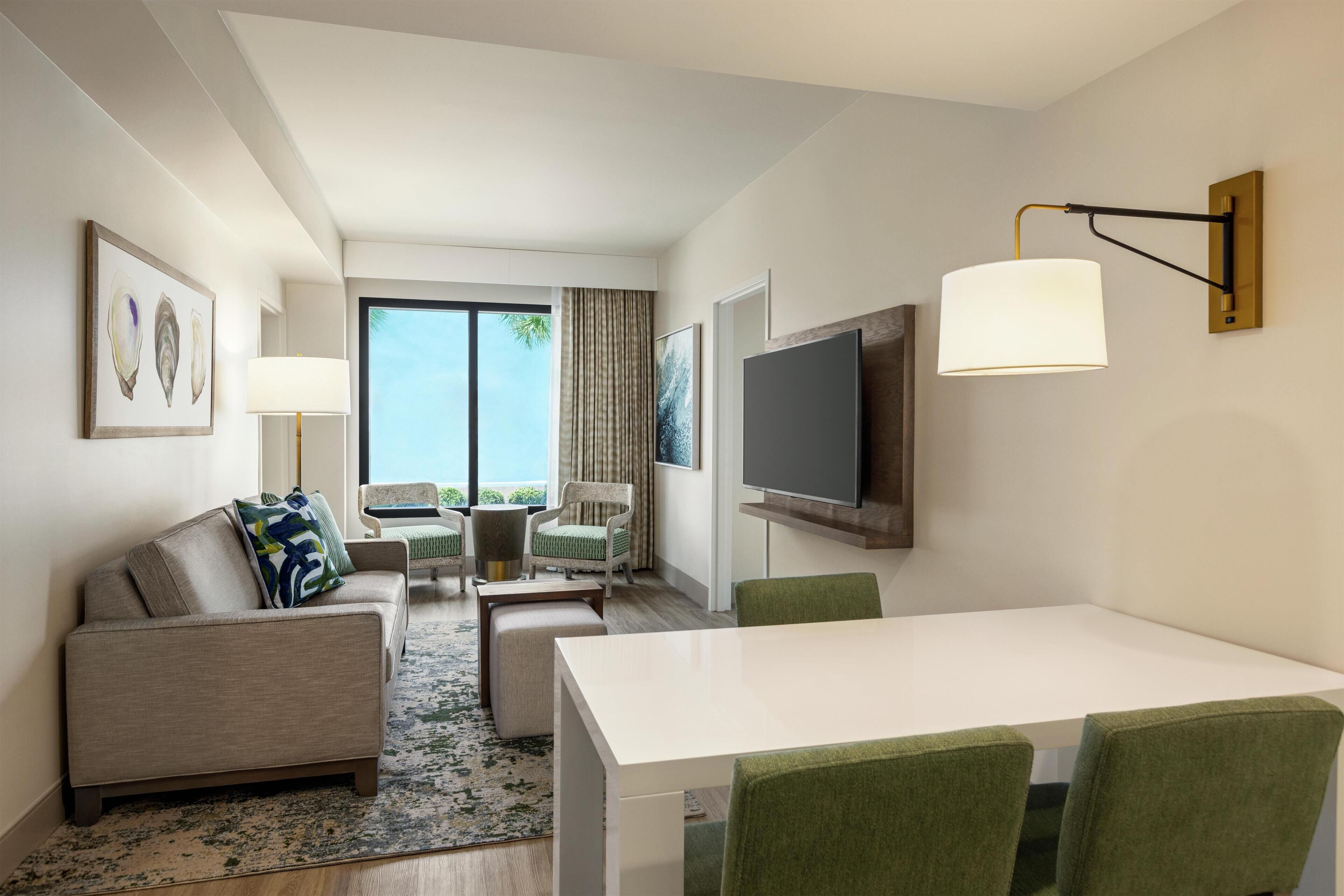 A modern hotel room with a sofa, chairs, a dining table, a TV, and a large window offering a view of the outdoors.