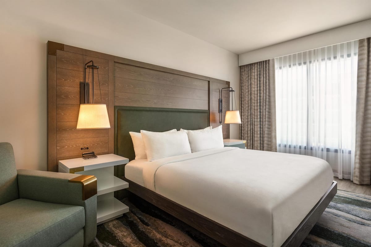 A hotel room with a large bed, two bedside tables with lamps, a green upholstered chair, and a view of the outdoors through sheer curtains.