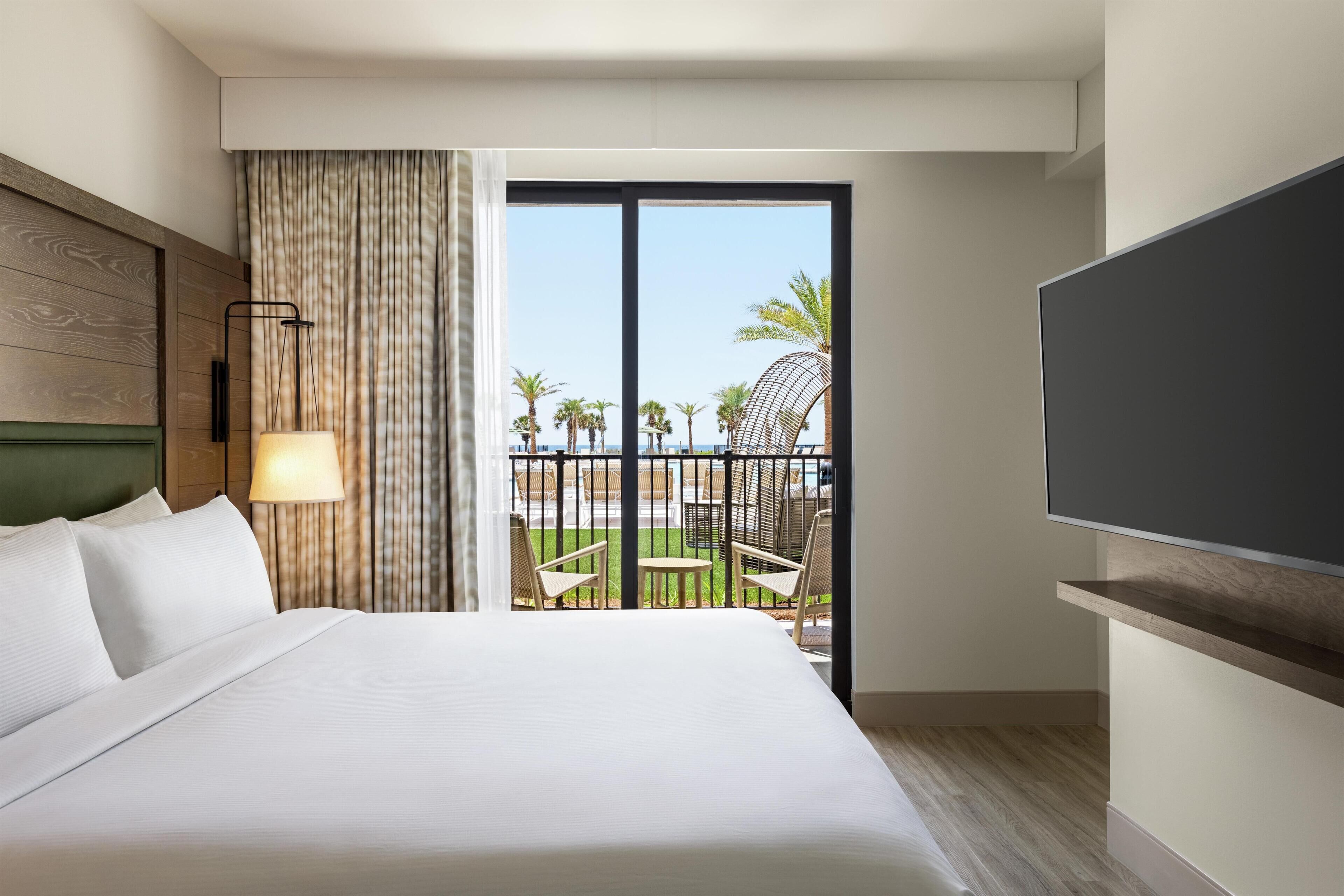 Quarto de hotel com uma cama grande, televisão, varanda com cadeiras e vista para a praia.