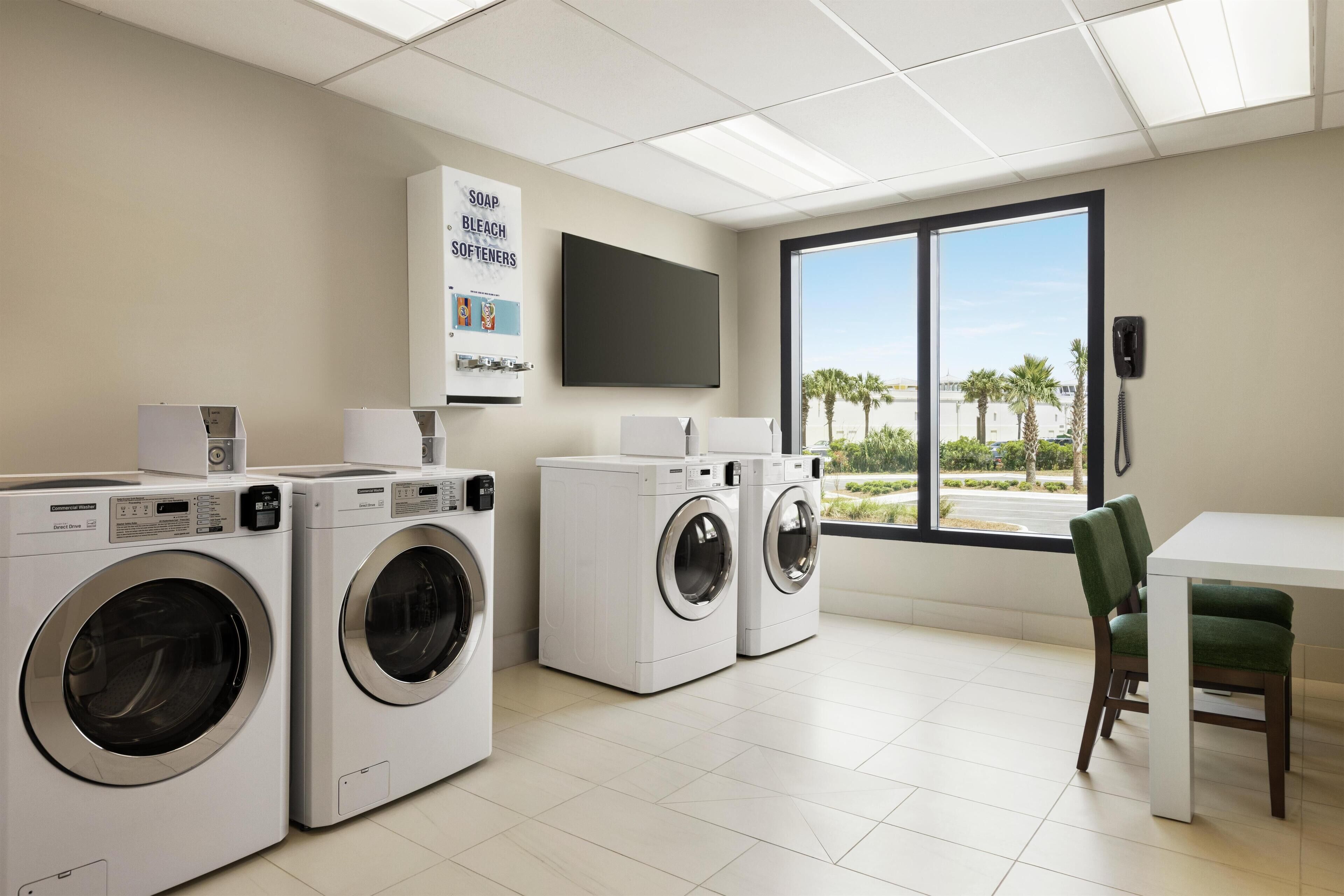 A laundry room with three washing machines, a large window with a view of palm trees, and a flat-screen TV mounted on the wall.