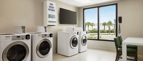 A laundry room with three washing machines, a large window with a view of palm trees, and a flat-screen TV mounted on the wall.