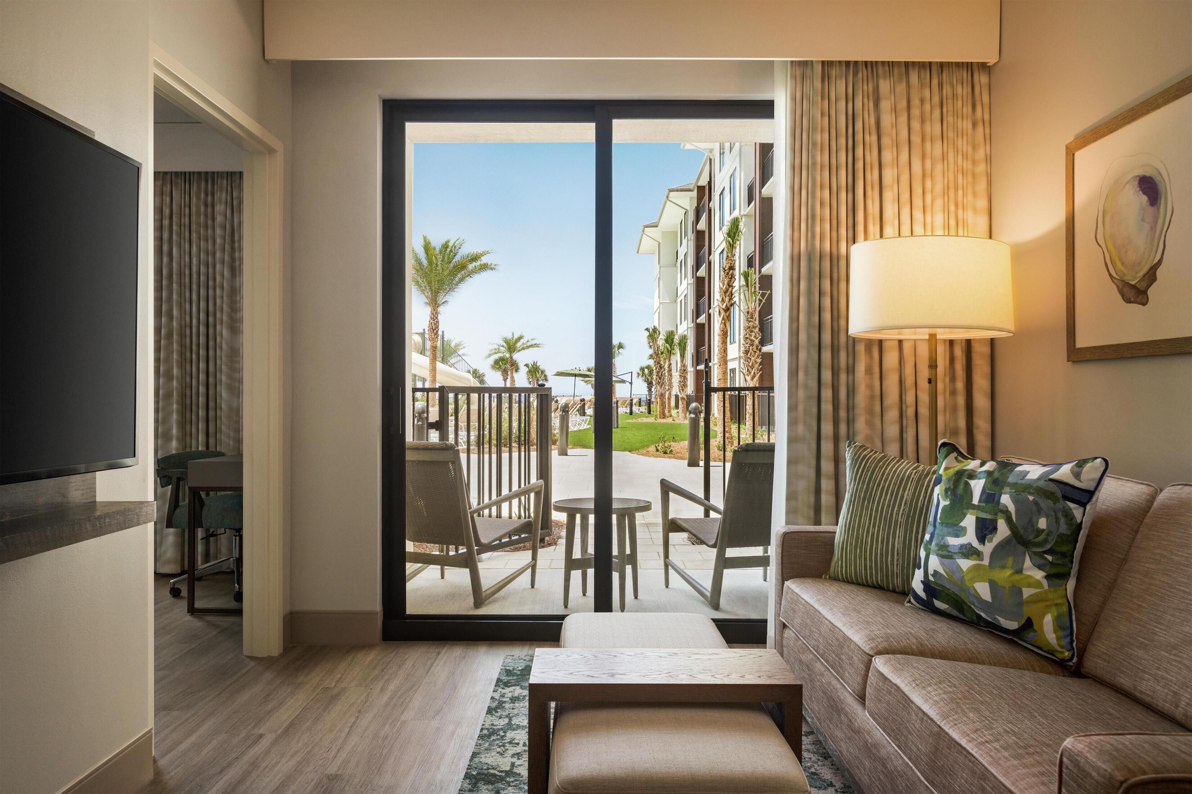 A hotel room with a sofa, a coffee table, a TV, and a sliding glass door leading to a balcony with outdoor seating and palm trees.