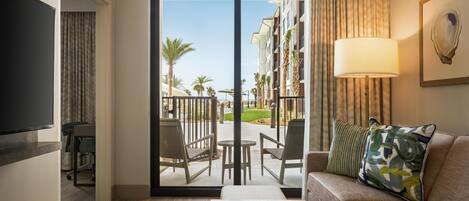 A hotel room with a sofa, a coffee table, a TV, and a sliding glass door leading to a balcony with outdoor seating and palm trees.