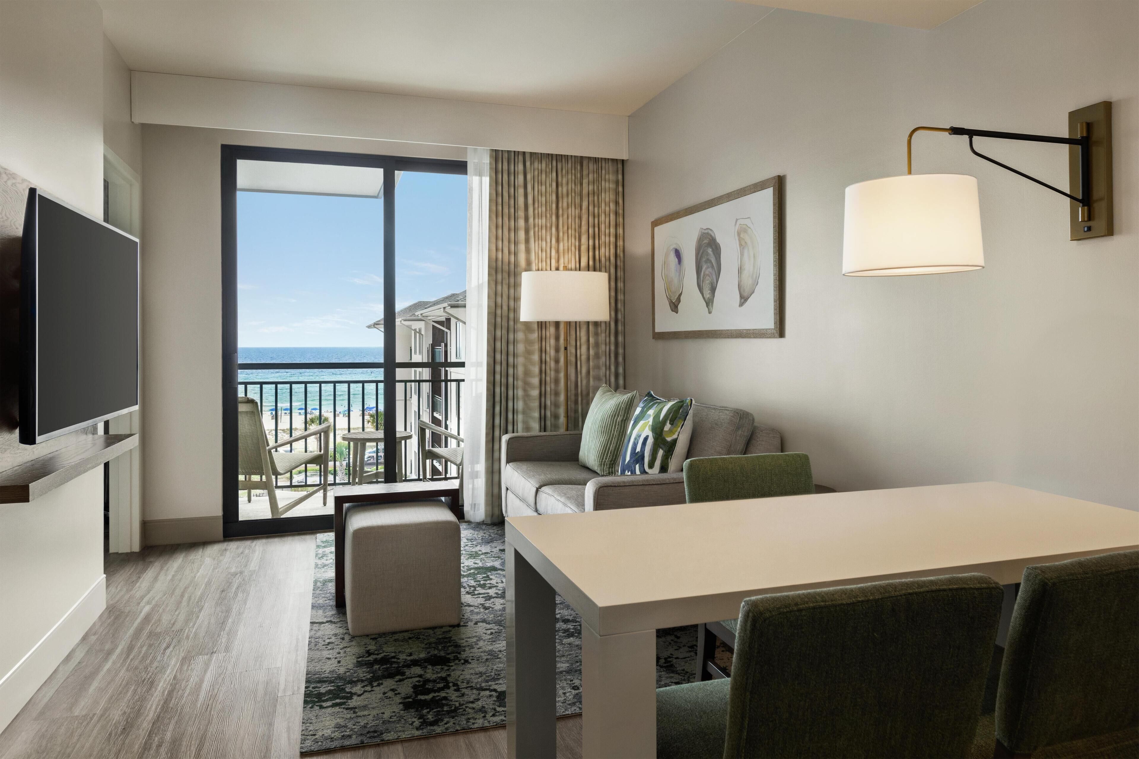 A hotel room with a balcony, a flat-screen TV, a dining table with chairs, a sofa with cushions, and a wall-mounted lamp.