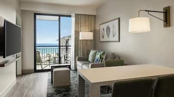 A hotel room with a balcony, a flat-screen TV, a dining table with chairs, a sofa with cushions, and a wall-mounted lamp.