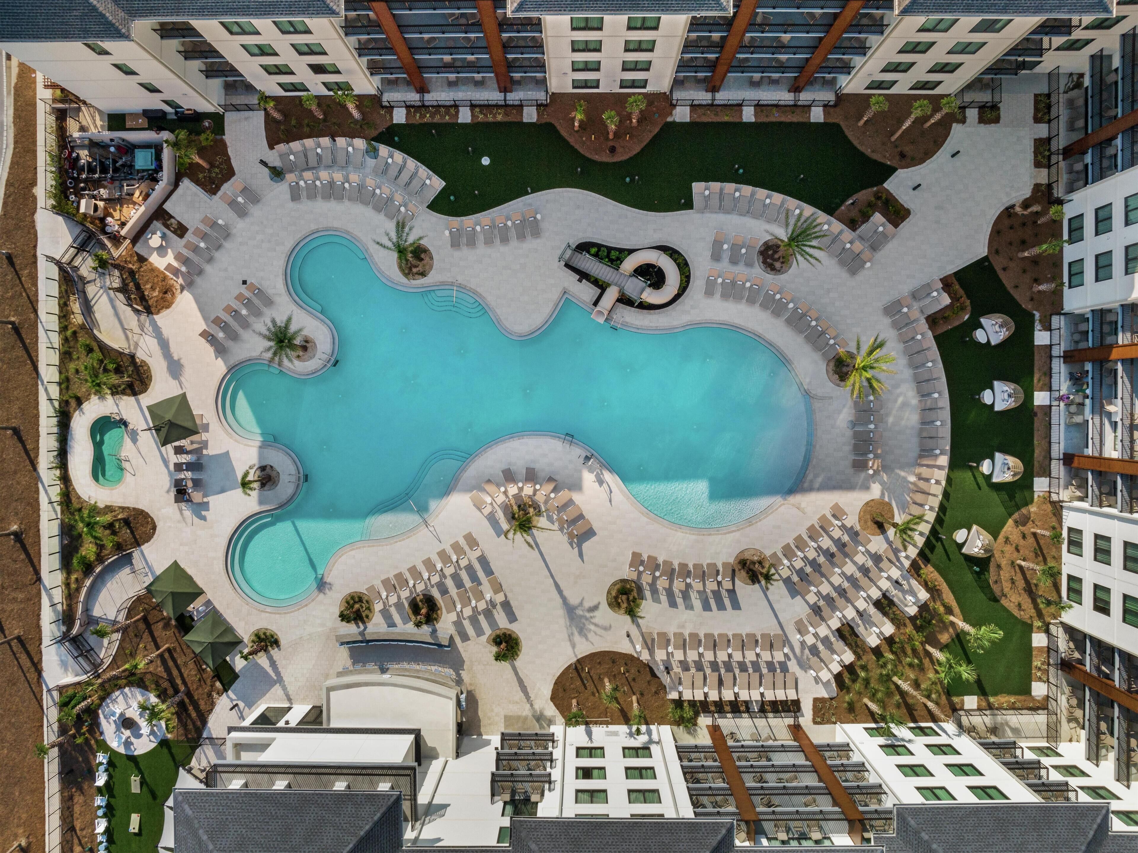 An aerial view of a hotel with a central swimming pool, multiple lounge areas with sunbeds, and a landscaped garden.