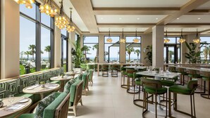 A restaurant with green upholstered booths and high tables, large windows with a view of the ocean, and pendant lighting. - Embassy Suites by Hilton Panama City Beach Resort (Panama City Beach)