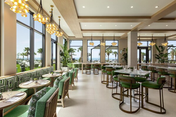 Un restaurante con asientos tapizados en verde y mesas altas, ventanas amplias con vistas al océano e iluminación colgante.