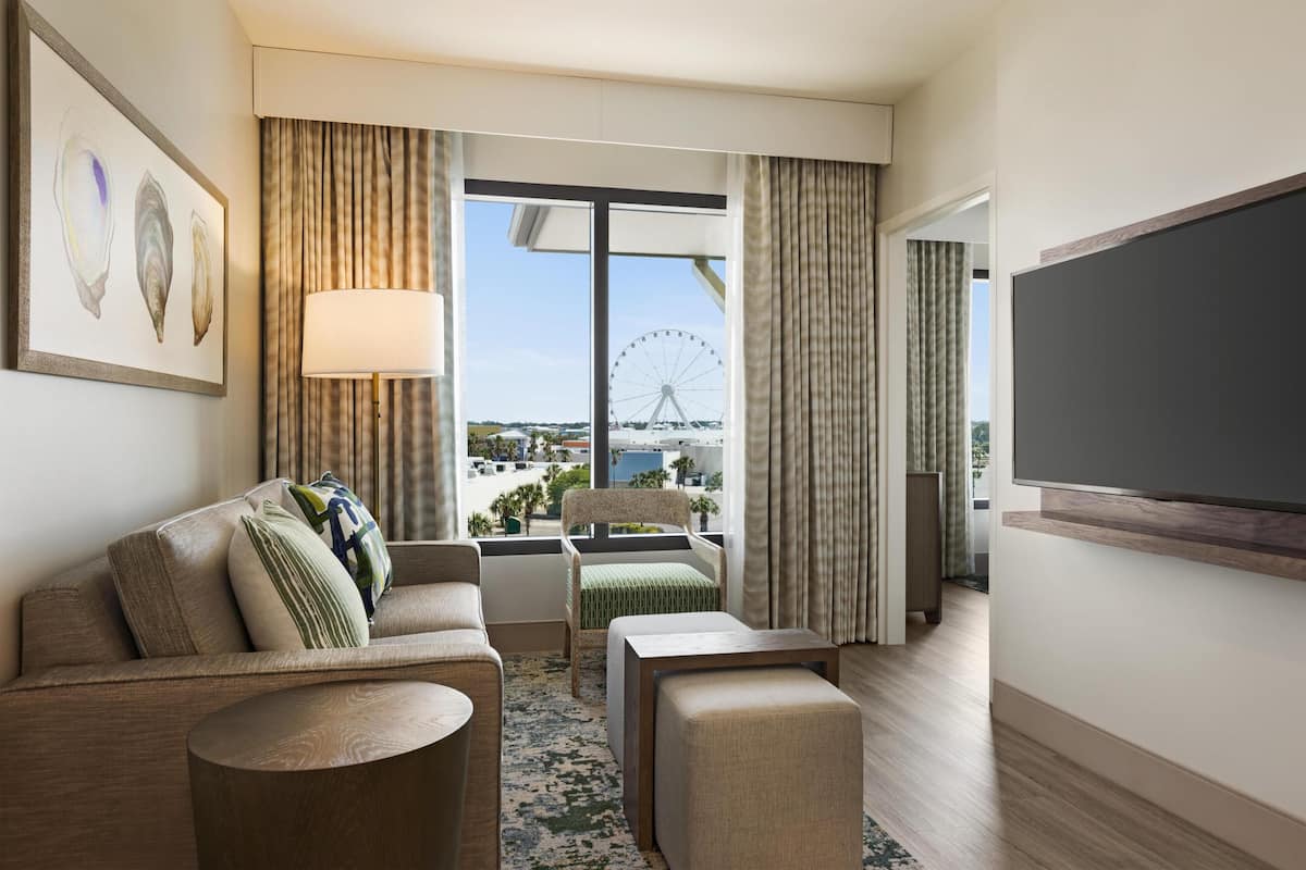 A hotel room with a large window, a sofa, a chair, a small table, and a flat-screen TV.