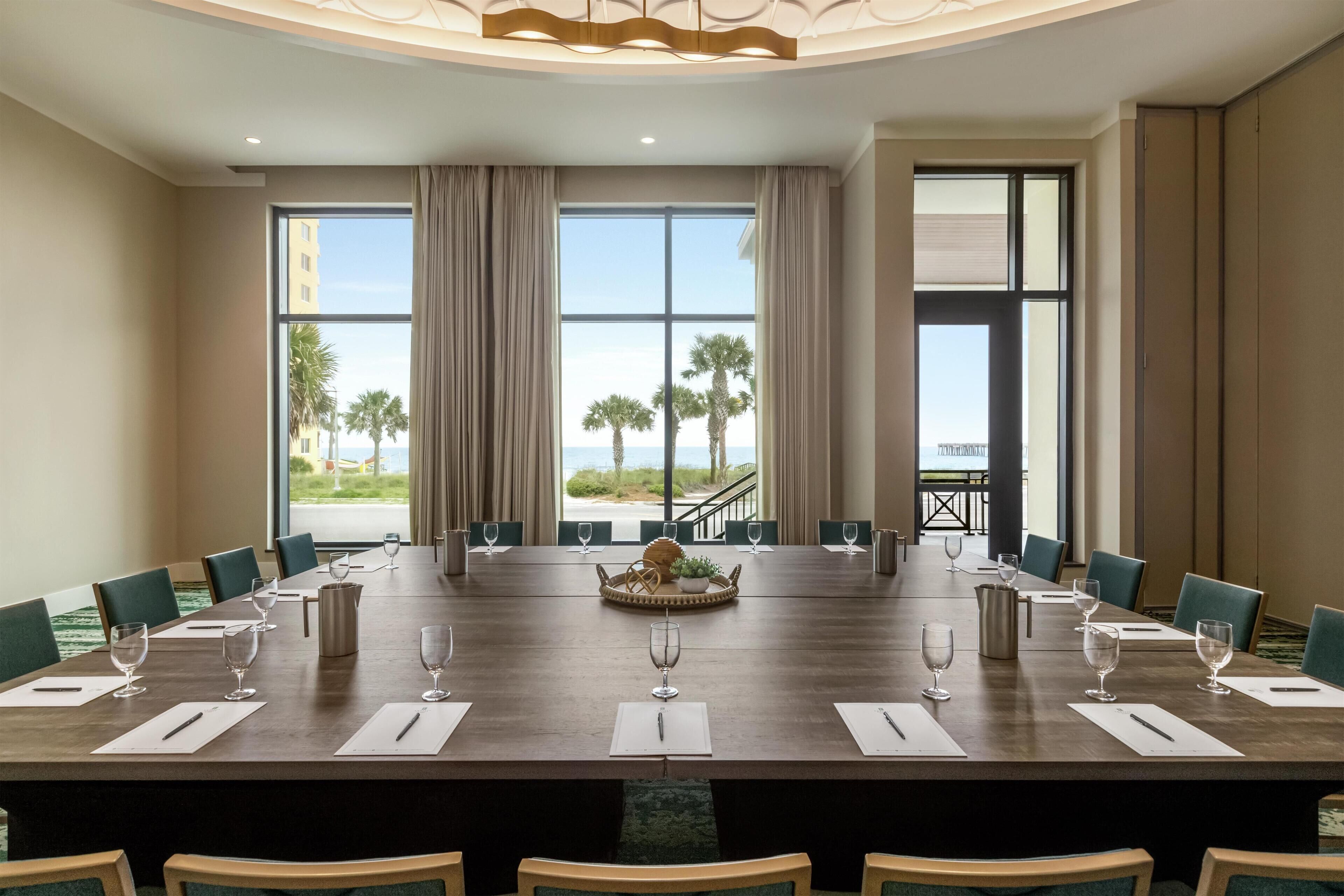 A conference room with a long table, chairs, and a view of palm trees and a staircase.