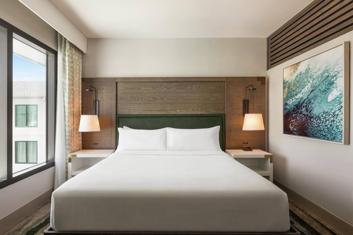 A hotel room with a large bed, two bedside tables with lamps, a window with curtains, and a large abstract painting on the wall.