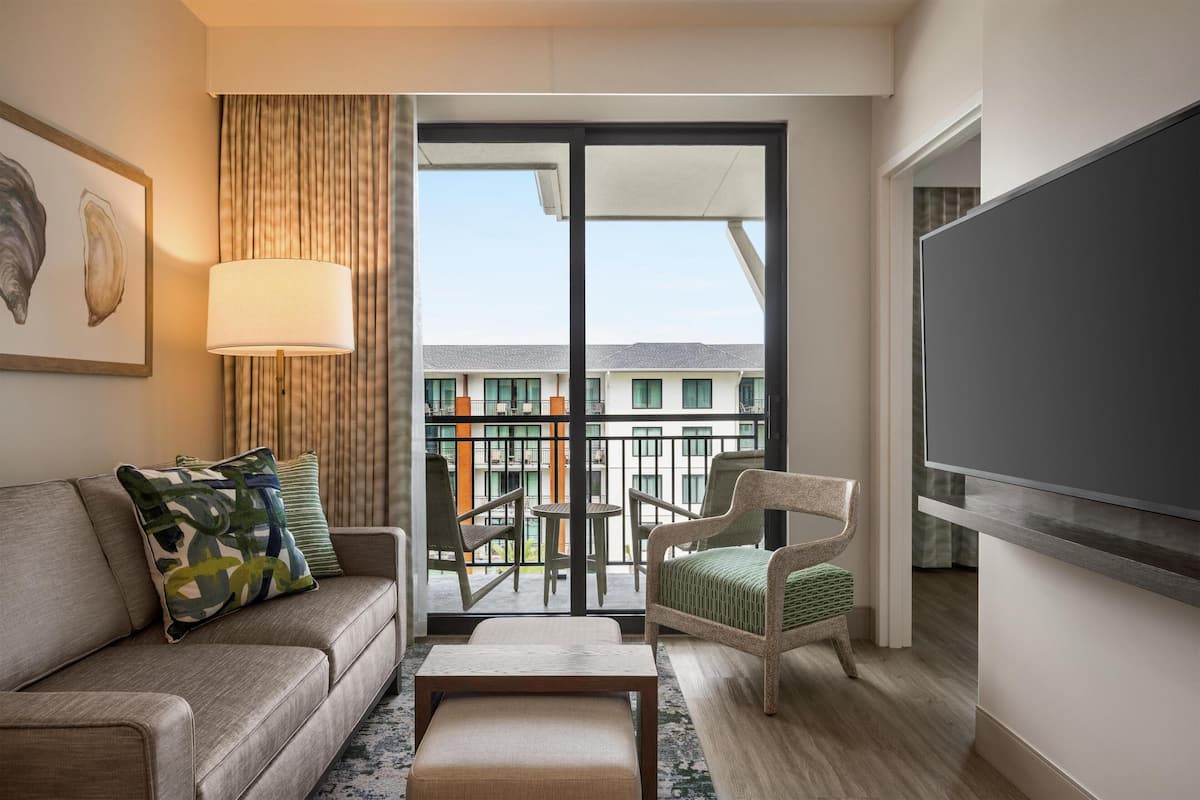 A hotel room with a sofa, armchair, and a table. There is a balcony with a view of buildings and a TV mounted on the wall.