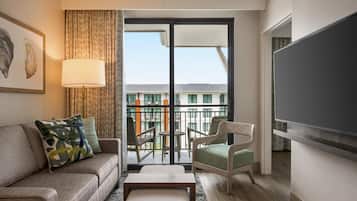 Habitación de hotel con sofá, sillón y una mesa. Tiene un balcón con vistas a edificios y un televisor montado en la pared.