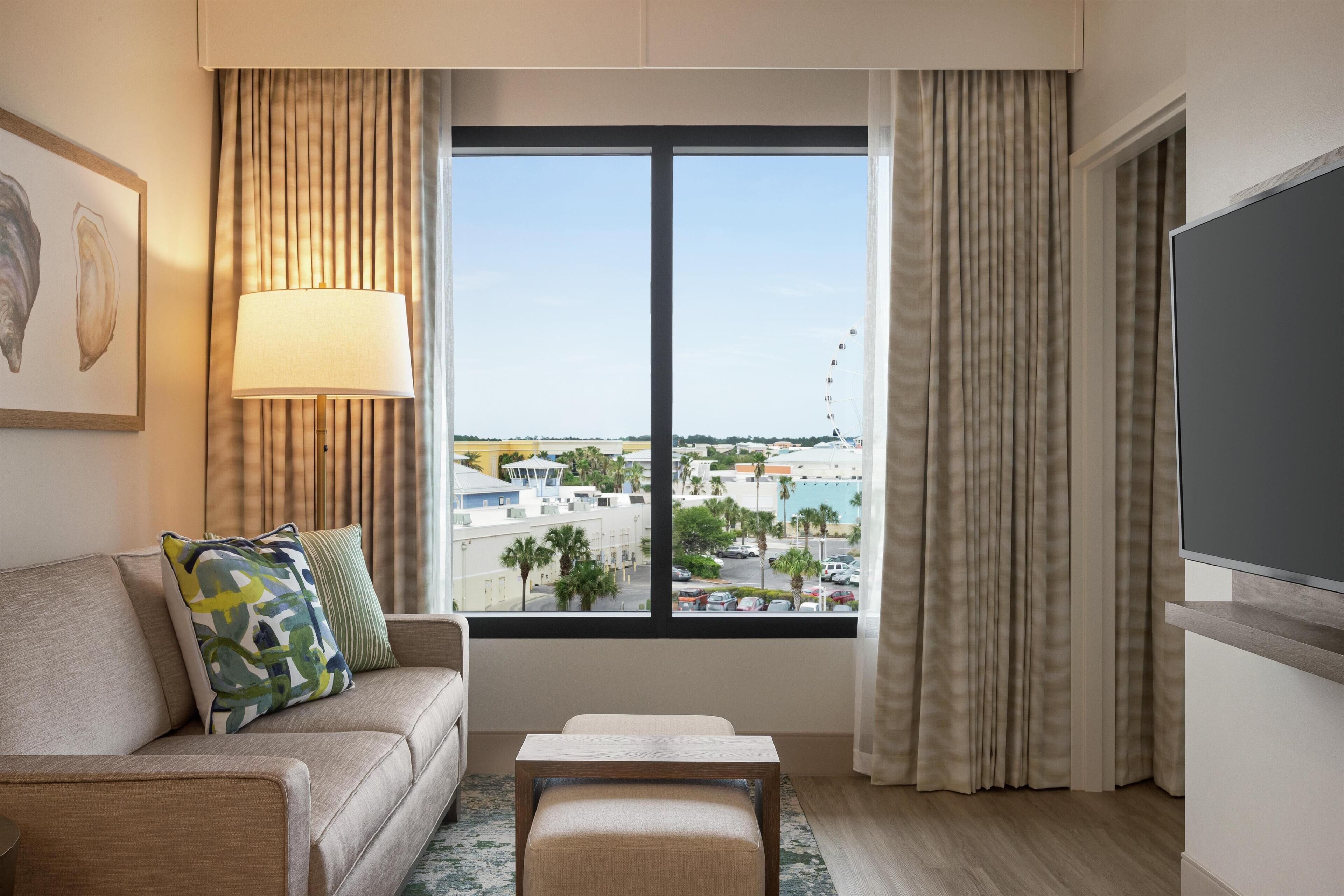 A hotel room with a sofa, a small table, a lamp, a TV, and a large window with a city view.