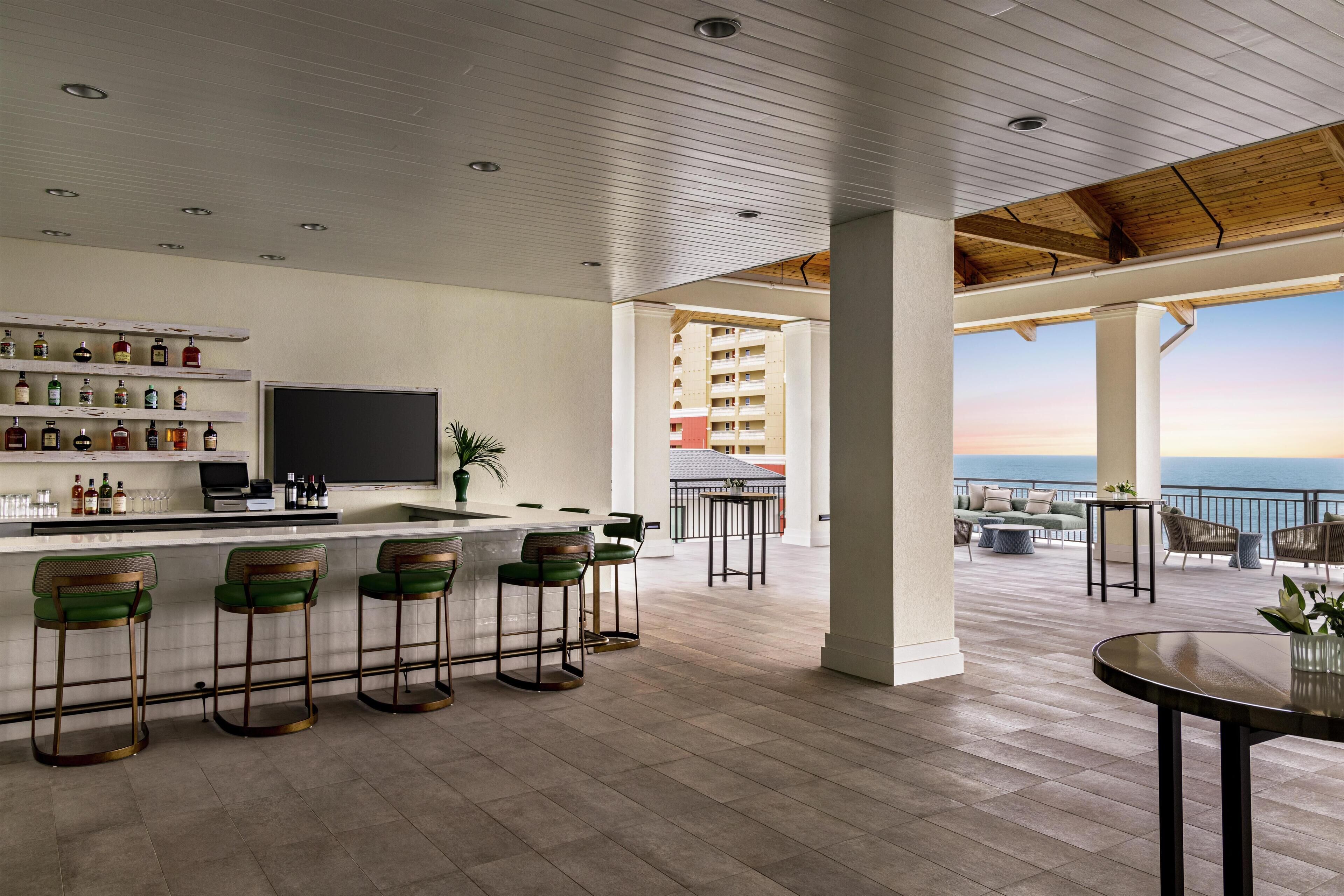 A bar area with stools, a counter, and shelves with bottles. There is a TV and a plant on the counter. The area has a view of the sea.