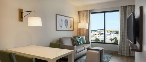 Ein modernes Hotelzimmer mit Essbereich, einer Sitzecke, einem Fernseher und einem großen Fenster mit Blick auf die Stadt.