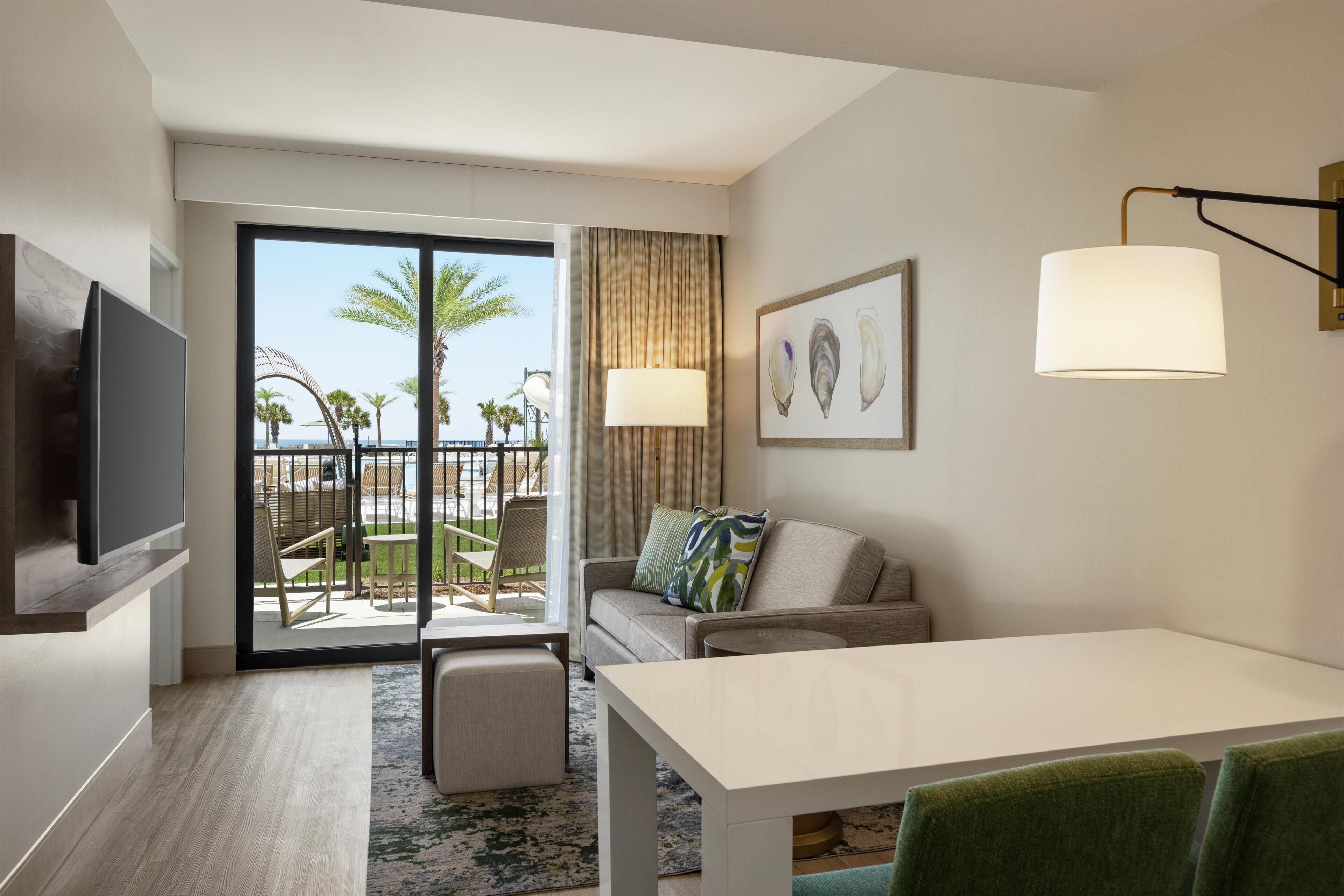 A modern hotel room with a large sliding glass door leading to a balcony with a view of palm trees and the ocean. There is a sofa, a small table, and a flat-screen TV mounted on the wall.