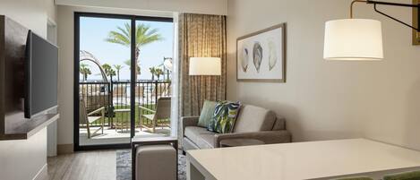 A modern hotel room with a large sliding glass door leading to a balcony with a view of palm trees and the ocean. There is a sofa, a small table, and a flat-screen TV mounted on the wall.