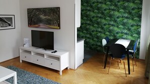 Una sala de estar moderna con una televisión de pantalla plana sobre un mueble blanco, un comedor con una mesa y sillas, y una pared con un diseño de hojas tropicales.
