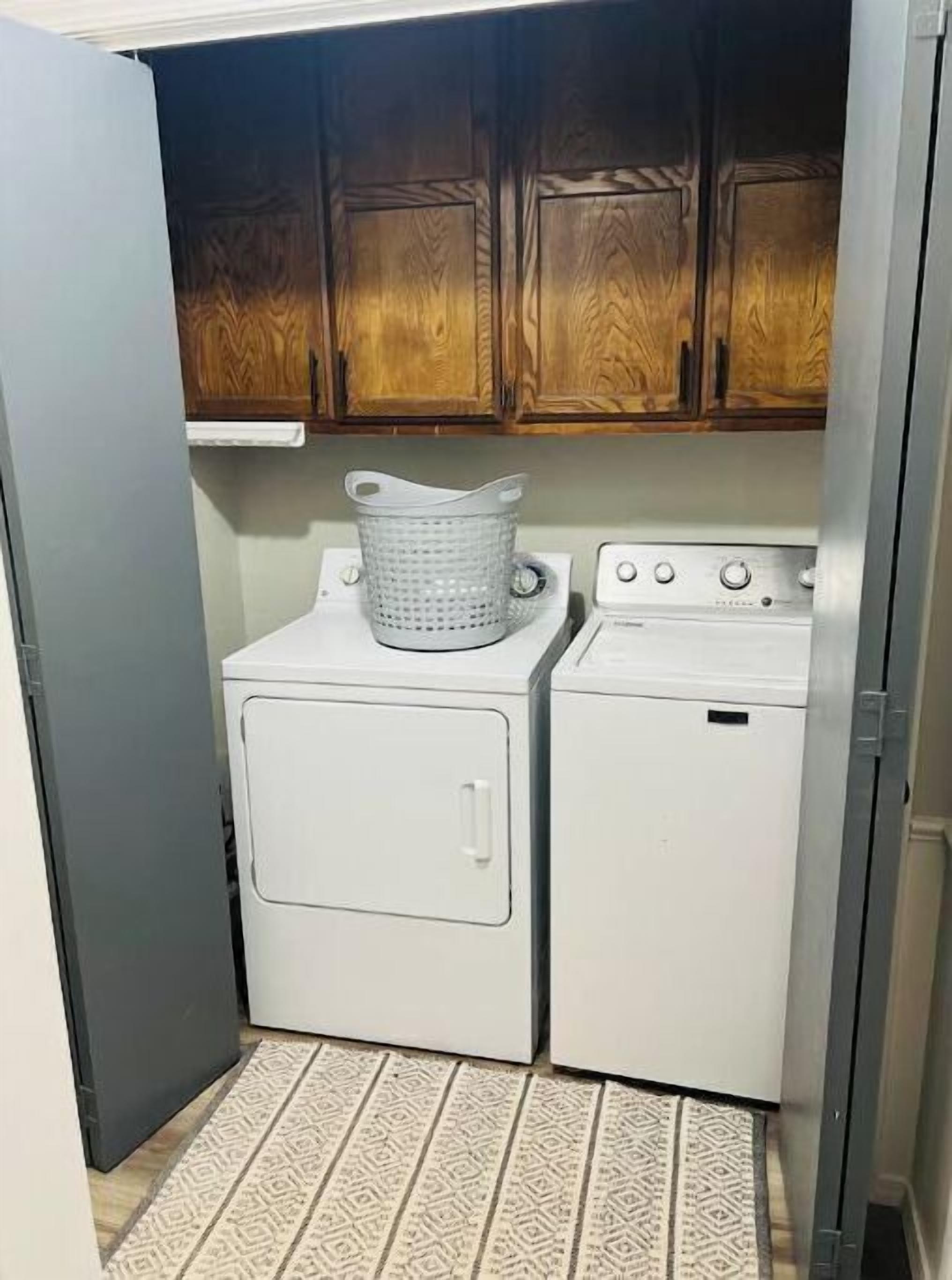 A compact laundry room with a front-loading washer and dryer, wooden cabinets above, and a patterned rug on the floor.