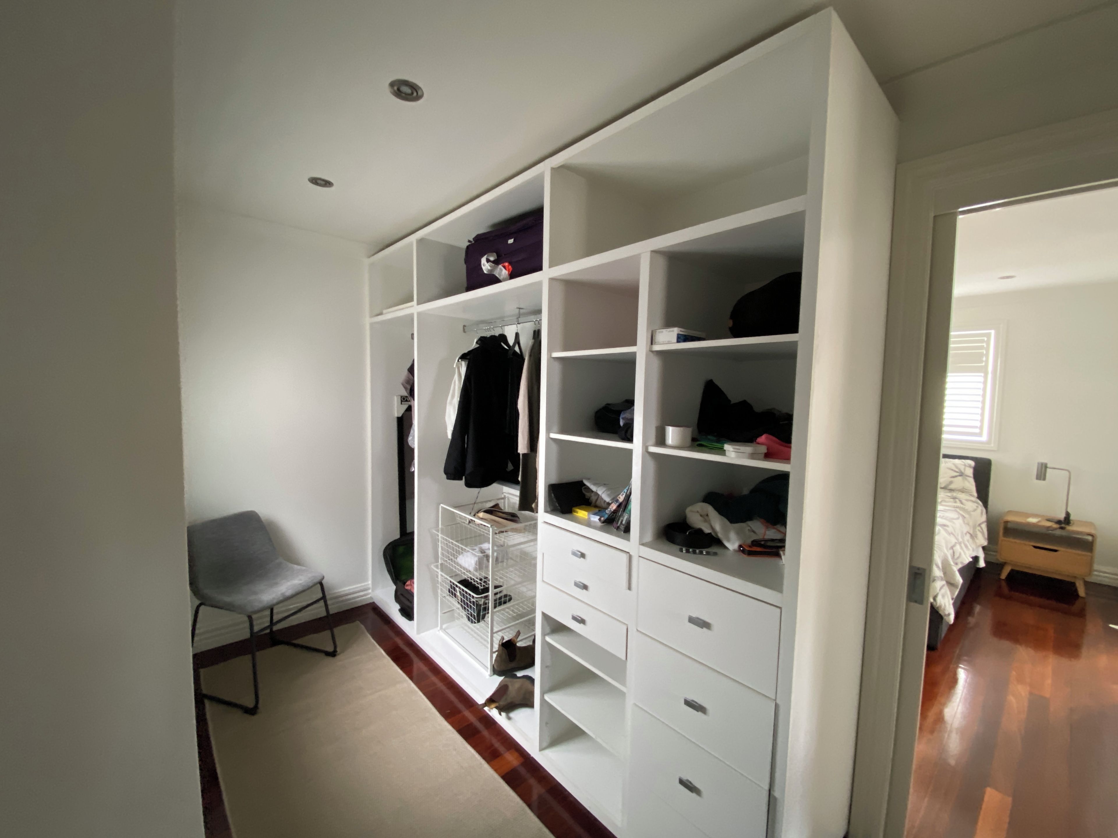 Un dressing avec de nombreuses étagères et des tiroirs, une chaise et un lit dans la pièce adjacente.