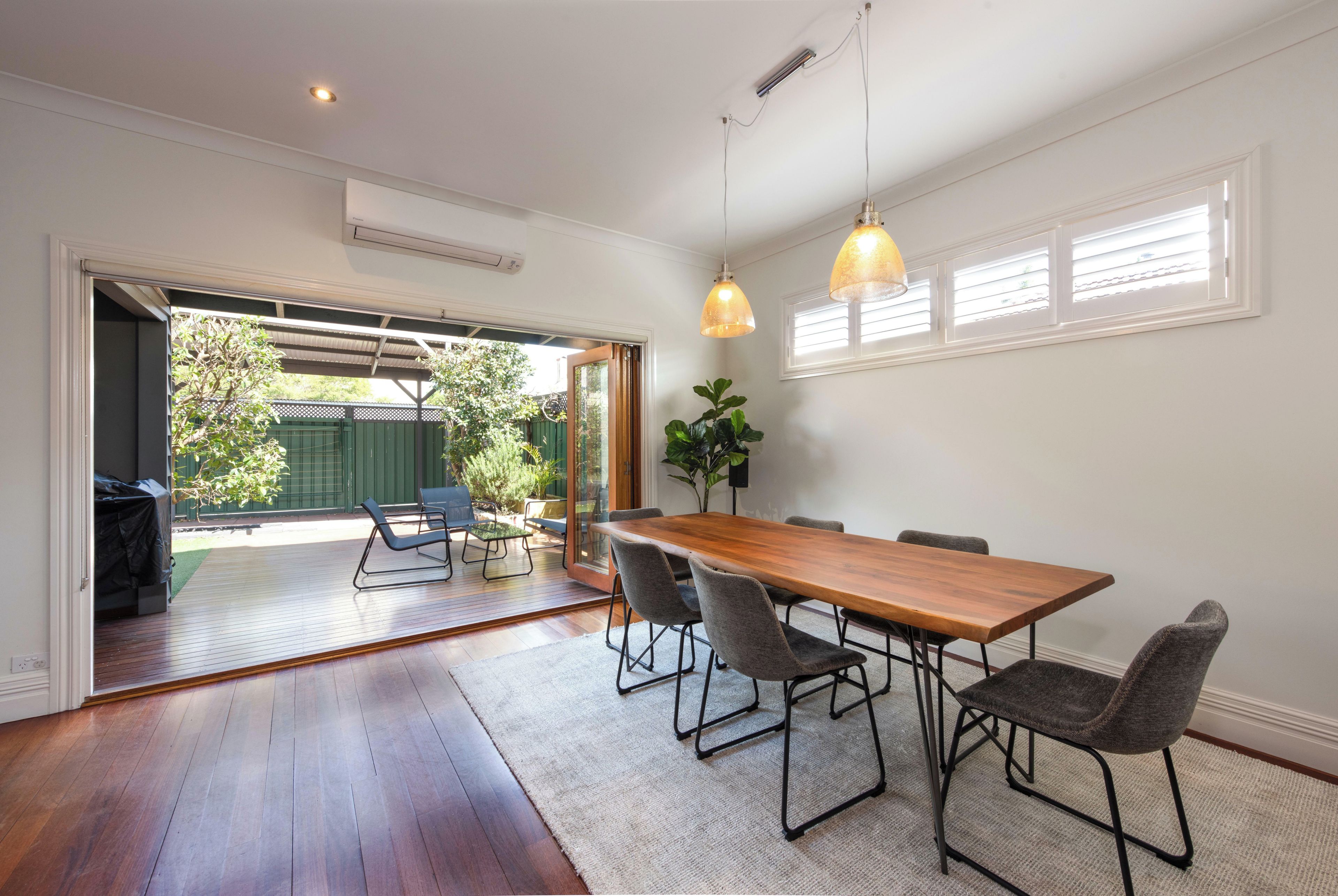 Un espace repas avec une table et des chaises en bois, une grande fenêtre avec des stores et une vue sur un espace extérieur verdoyant.