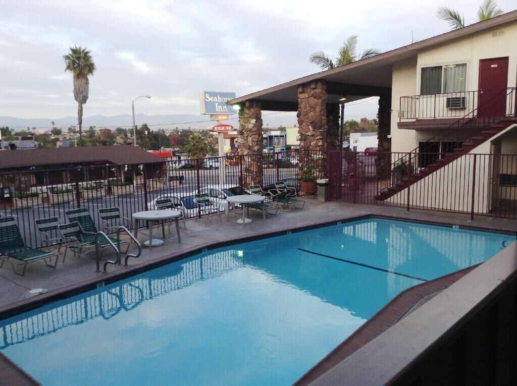 Una piscina con sillas de descanso y mesas, un área de estar techada y vista a un motel de fondo.