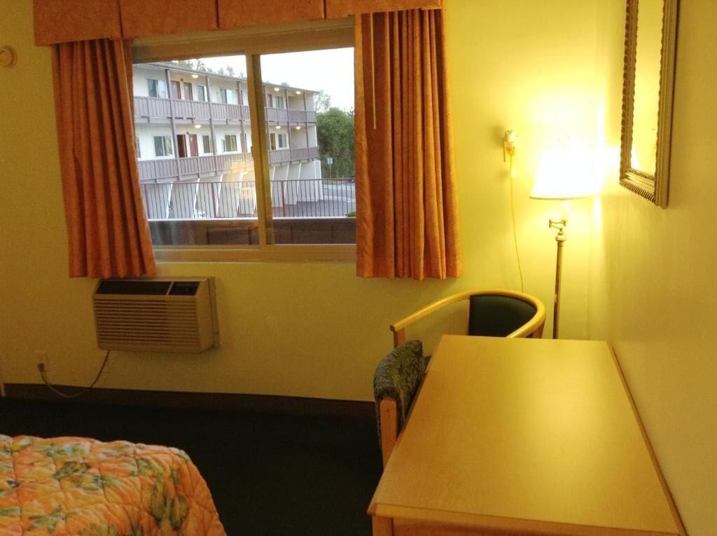 A hotel room with a bed, a desk, a chair, a window with curtains, an air conditioning unit, and a lamp.