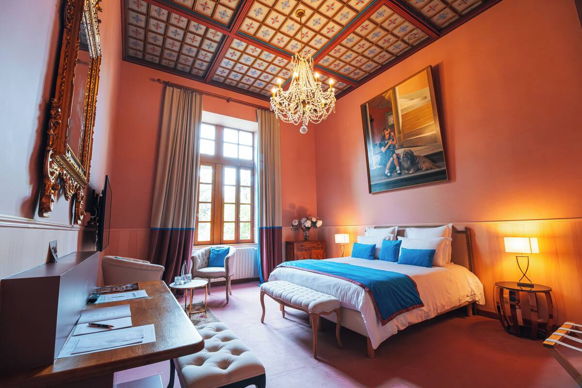 A hotel room with a large bed, a desk with papers, a chair, and a chandelier.