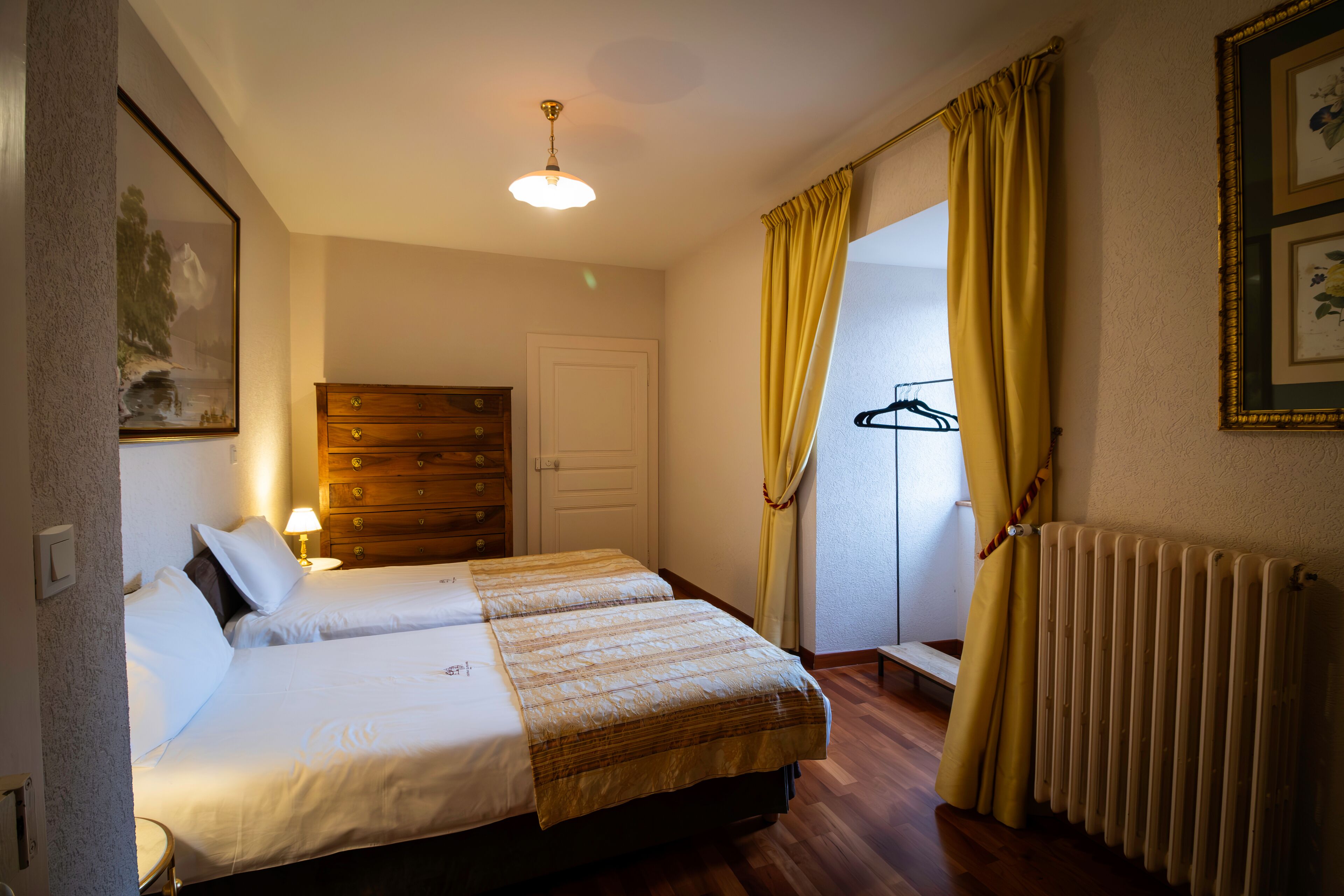 A hotel room with two beds, a wooden dresser, a radiator, and a window with yellow curtains.
