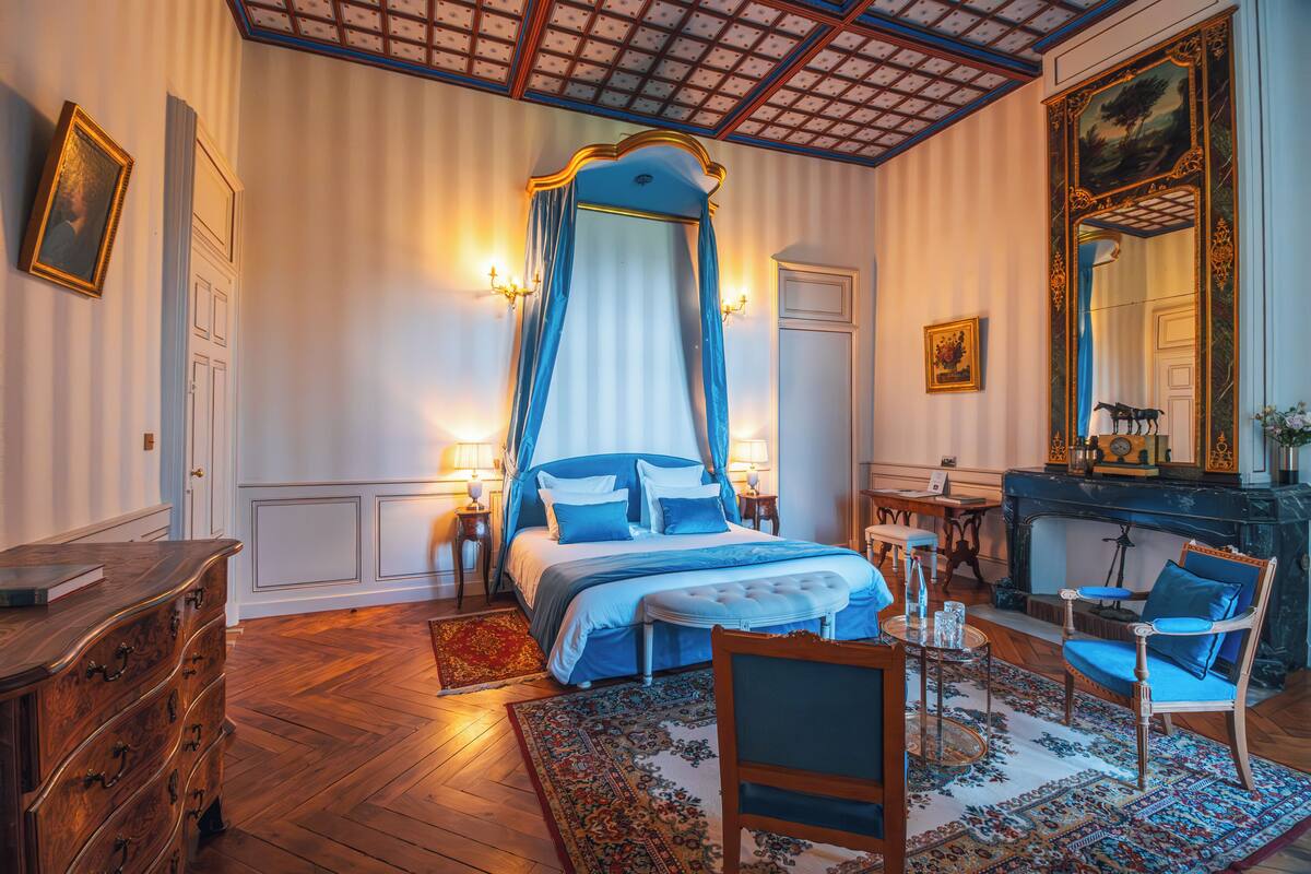 A bedroom with a four-poster bed, a sitting area with a chair and table, and a fireplace.