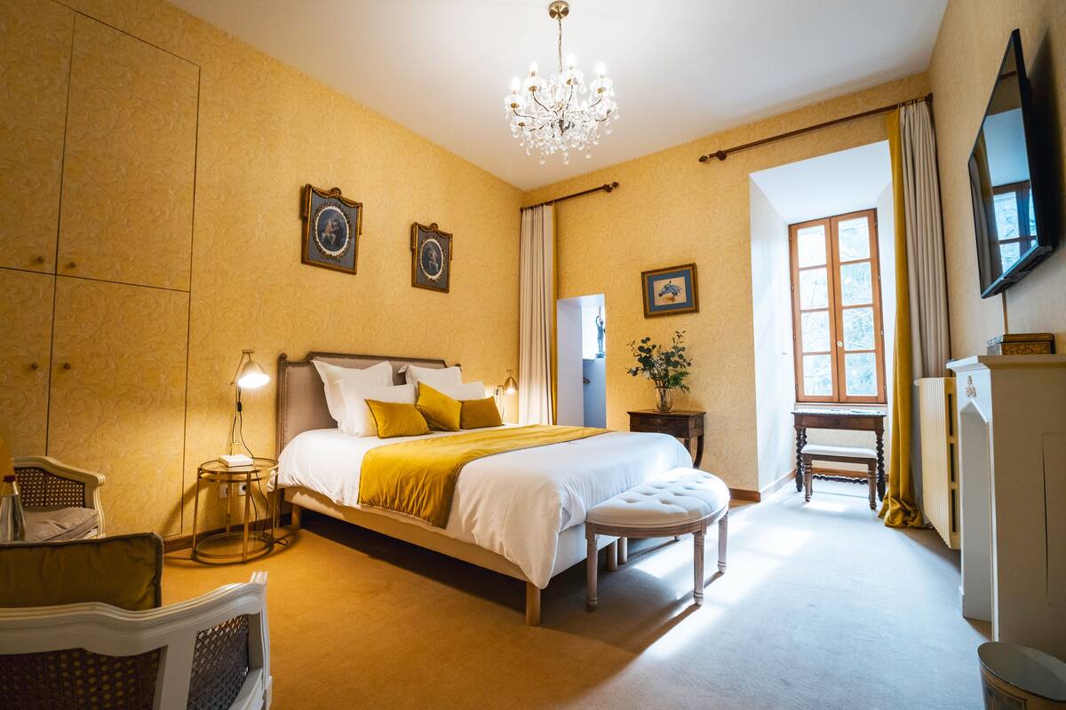 A hotel room with a large bed, a chandelier, a sitting area with a chair and table, and a television mounted on the wall.