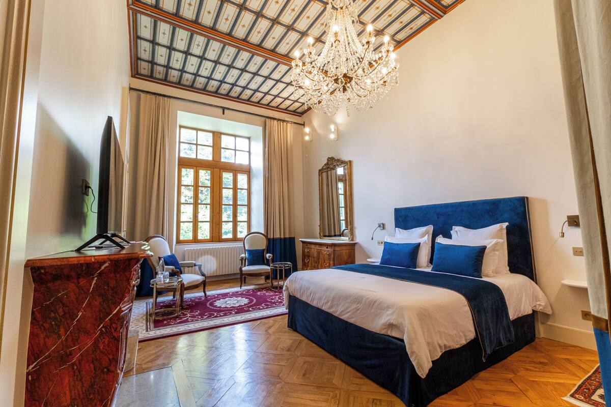 A spacious bedroom with a large bed, a wooden desk, a chandelier, and a large window with curtains.
