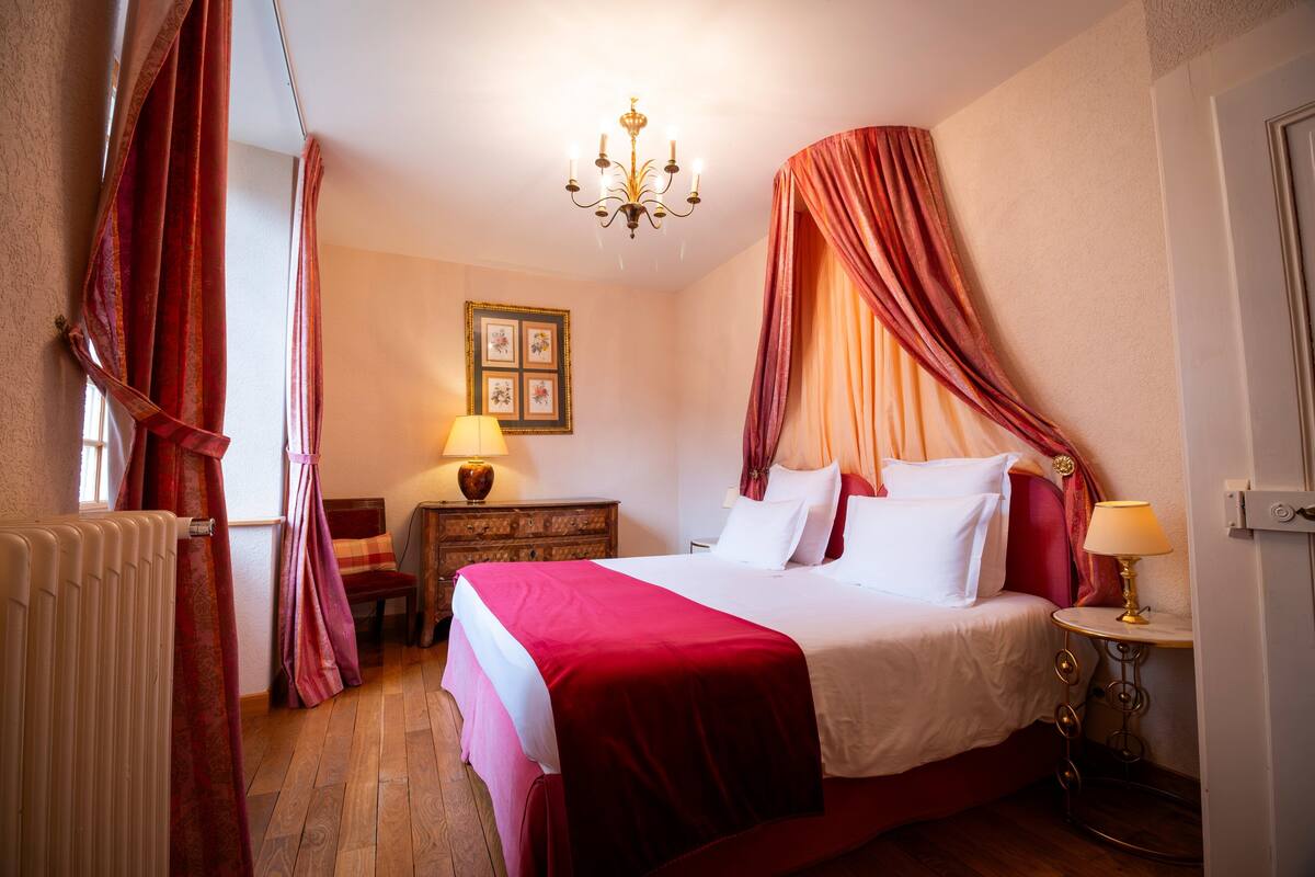 A bedroom with a four-poster bed, a chandelier, a wooden nightstand, and a small table with a lamp.