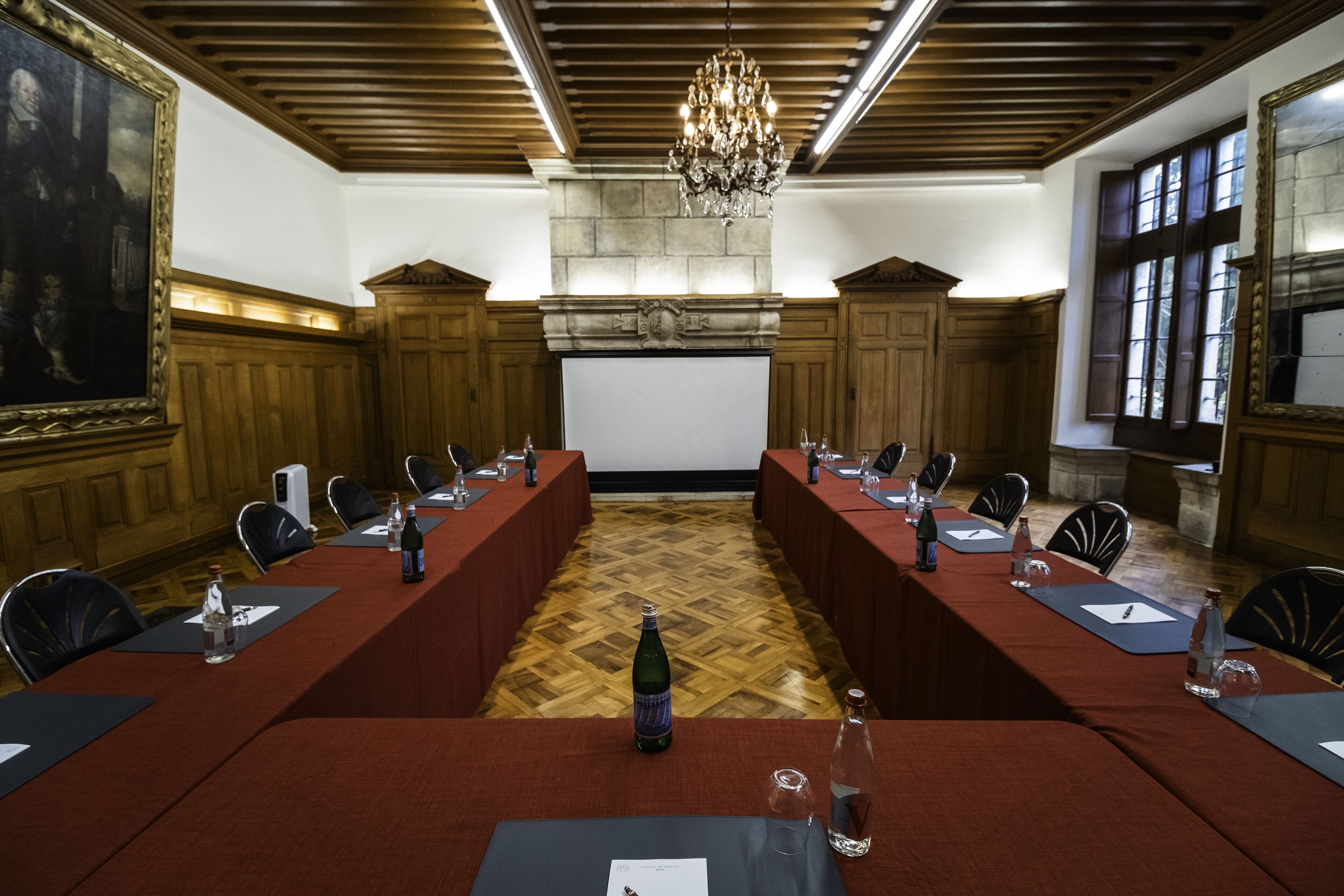 Μια αίθουσα συνεδριάσεων με ένα μεγάλο τραπέζι, καρέκλες, μια οθόνη προβολής και ένα πολυέλαιο.