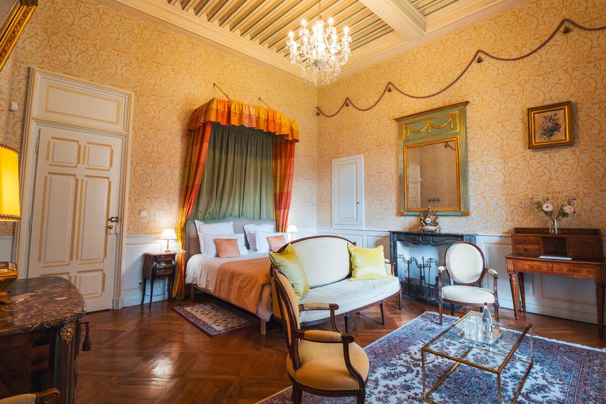 A hotel room with a bed, two chairs, a small table, a mirror, and a chandelier.