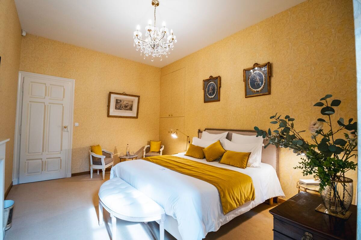 A hotel room with a bed, a desk, a chair, a chandelier, and two framed pictures on the wall.