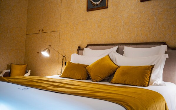 A neatly made bed with white and mustard-colored linens, a bedside lamp, and a wall with a floral pattern. - Château de Thorens (Fillière)