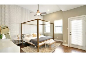 A bedroom with a four-poster bed, a bedside table with a lamp, a round mirror, and a window with blinds.